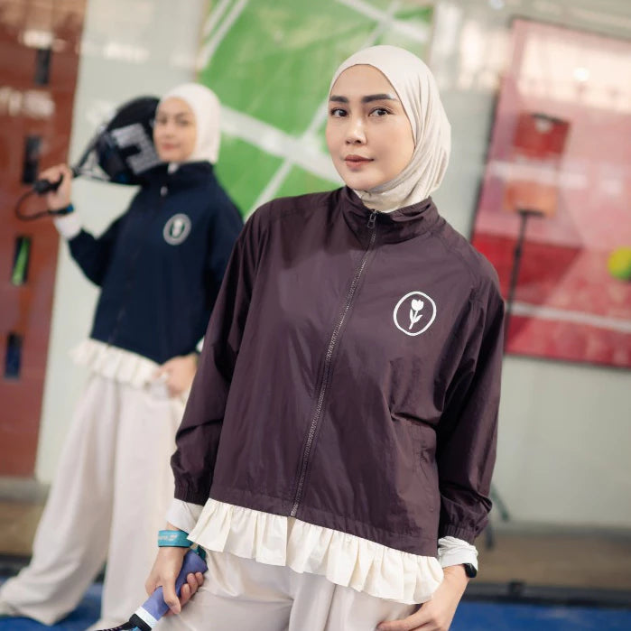 A woman wearing a white hijab and a dark plum zip-up jacket by Haidee Orlin with a white ruffled hem stands in an indoor sports facility. Another person is blurred in the background holding a camera.