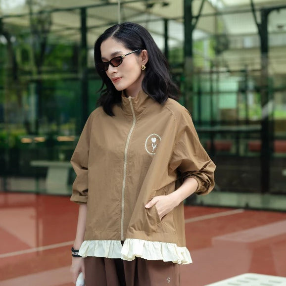 A woman wearing a brown lightweight zip-up jacket by Haidee Orlin with a white ruffled hem and a small circular chest graphic, standing in an outdoor modern architectural setting.