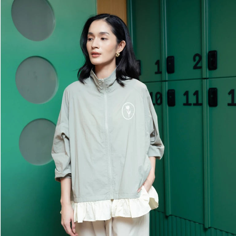 A woman wearing a light sage green zip-up jacket by Haidee Orlin with a white circular logo on the chest, layered over a cream ruffled top and cream pants, standing in front of green metal lockers.