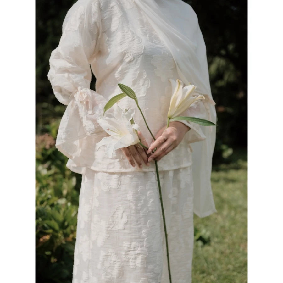 Person wearing a broken white lace blouse with floral embroidery by Renaette holding a flower in a natural setting