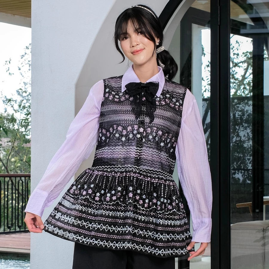 Woman wearing a black patterned outer by Holy Peony over a light purple shirt outdoors.