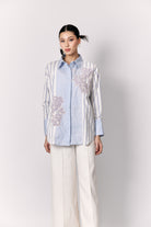 Woman wearing a light blue and white striped shirt with floral embroidery by Studio Tui on a white background