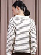 A woman shown from the back wearing a cream-colored long-sleeve polo shirt by Hijabchic with thin brown horizontal stripes, set against a soft beige curtain background.