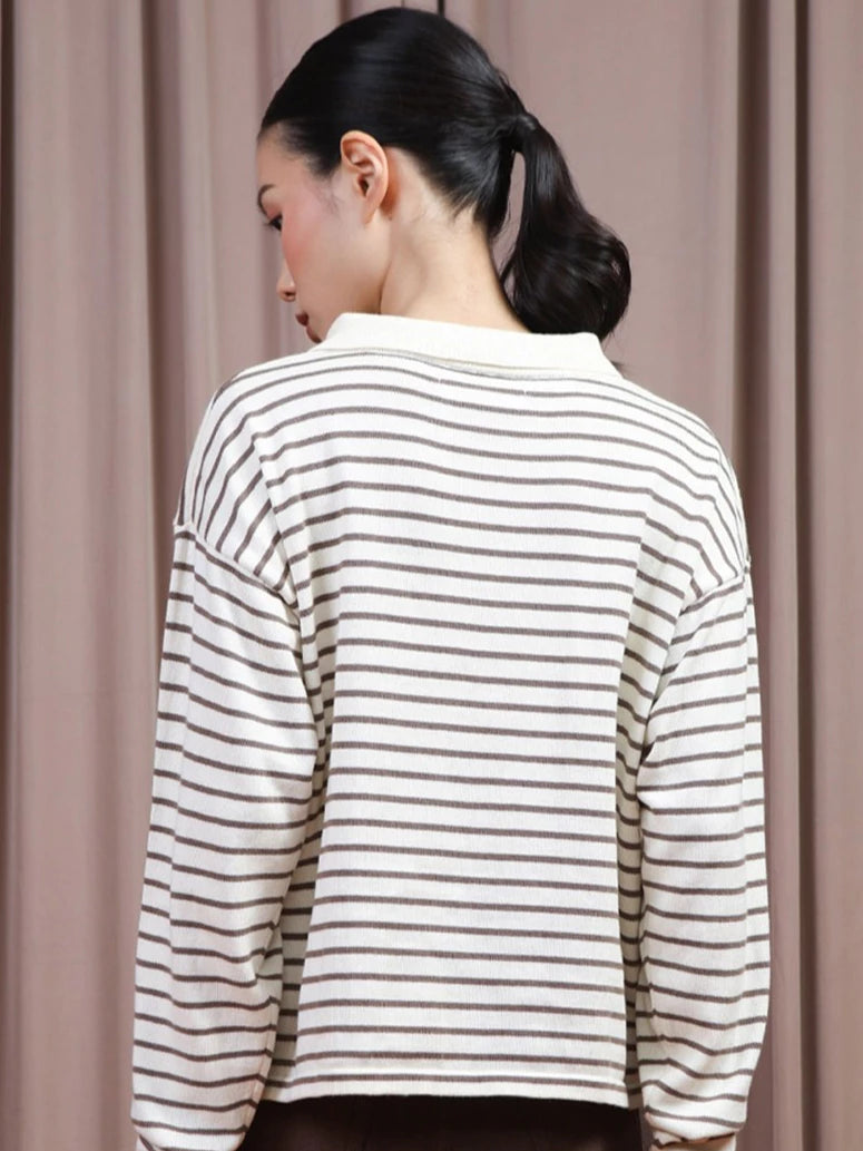 A woman shown from the back wearing a cream-colored long-sleeve polo shirt by Hijabchic with thin brown horizontal stripes, set against a soft beige curtain background.