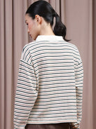 A woman shown from the back wearing a long-sleeve striped polo shirt by Hijabchic with thin horizontal green, black, and beige stripes on an off-white base, set against a soft beige curtain background.