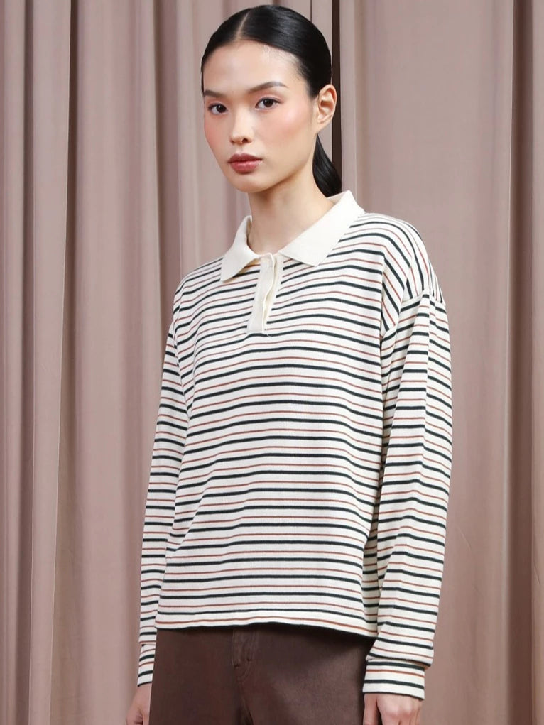 A model wearing a long-sleeve polo shirt by Hijabchic with thin horizontal green and beige stripes on an off-white base, paired with brown trousers, set against a backdrop of draped beige curtains.