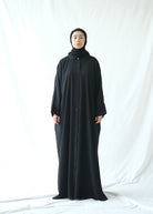 A person wearing a black traditional abaya by Rashawl with a button opening on the front, standing against a white background.