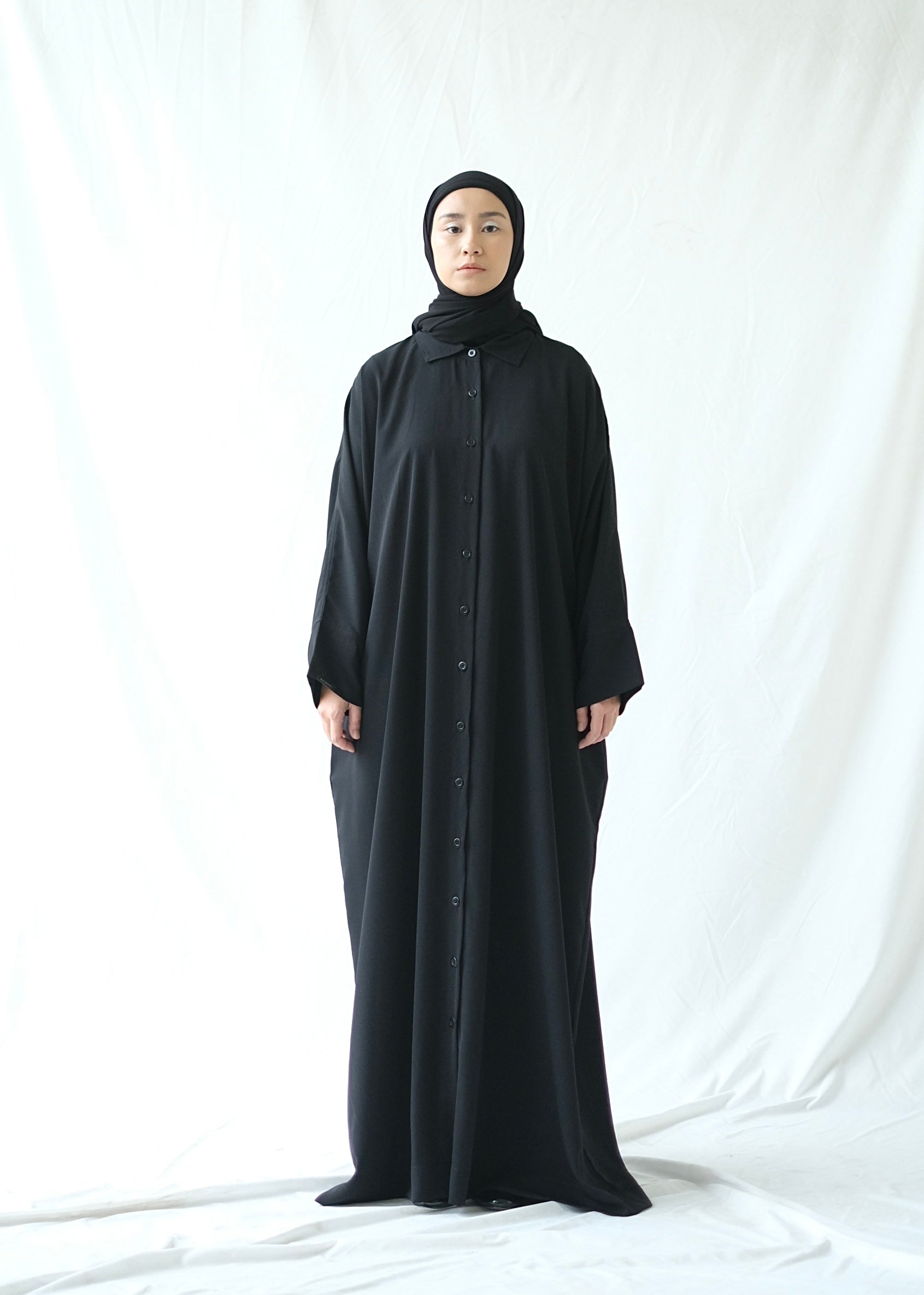 A person wearing a black traditional abaya by Rashawl with a button opening on the front, standing against a white background.