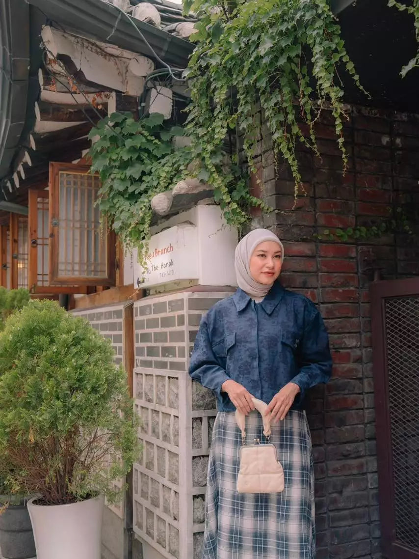 A woman wearing a blue floral long-sleeve outer by Haidee Orlin and a blue and white plaid skirt, paired with a light beige hijab and holding a small beige handbag, standing in front of a brick wall with green ivy in a courtyard setting.