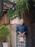 A woman wearing a blue floral long-sleeve outer by Haidee Orlin and a blue and white plaid skirt, paired with a light beige hijab and holding a small beige handbag, standing in front of a brick wall with green ivy in a courtyard setting.