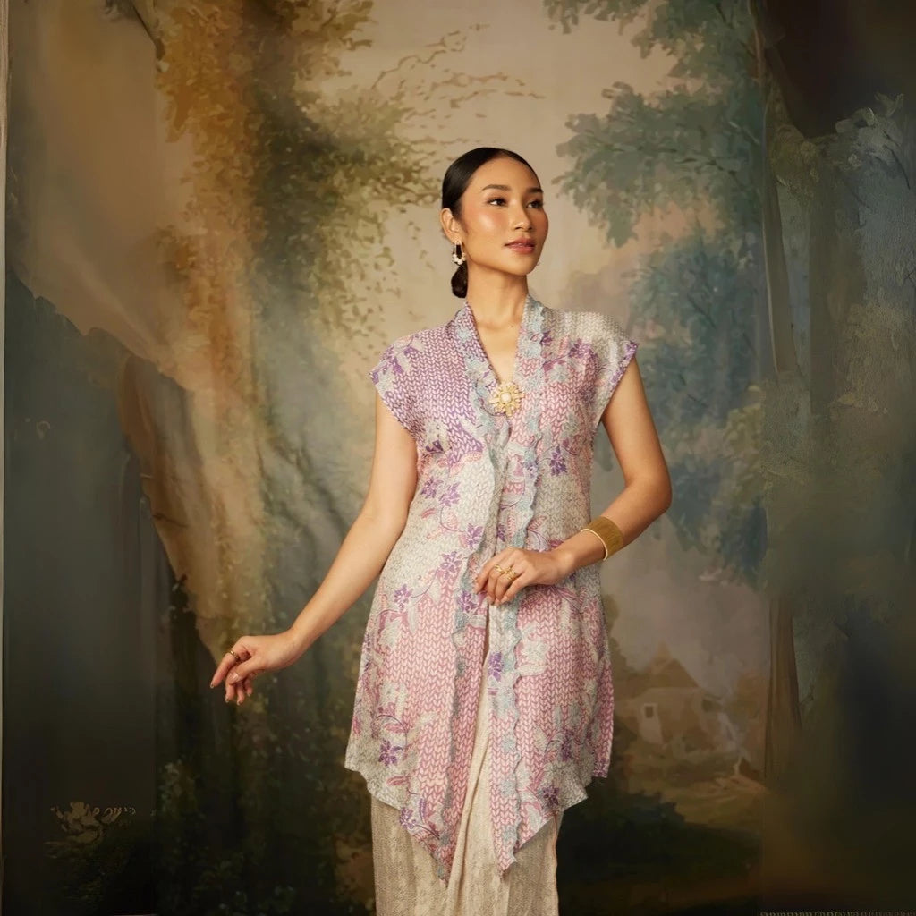 A woman wearing a lavender colored, long-sleeved velvet top by Kartinis Label with detail pita and manik, standing in front of a painted backdrop.