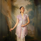 A woman wearing a lavender colored, long-sleeved velvet top by Kartinis Label with detail pita and manik, standing in front of a painted backdrop.