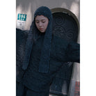 A person wearing a dark charcoal gray textured top by Yestoday, including a crocheted beanie with a long neck drape, standing in front of a building door.