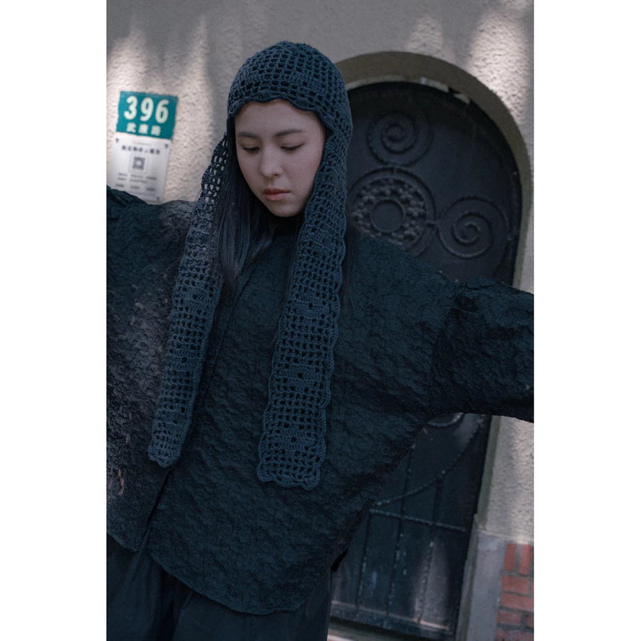 A person wearing a dark charcoal gray textured top by Yestoday, including a crocheted beanie with a long neck drape, standing in front of a building door.