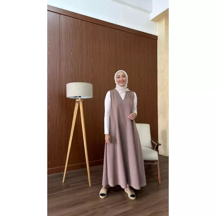 Woman in a brown long sleeveless dress by Abame standing in a room with wooden paneling and a lamp.