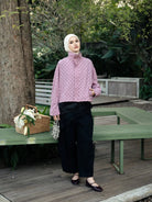 A woman wearing a light pink long-sleeve turtleneck shirt by Haidee Orlin with a small black polka dot pattern, paired with black wide-leg pants, a white hijab, and dark flat shoes, standing on a wooden deck in a garden setting.