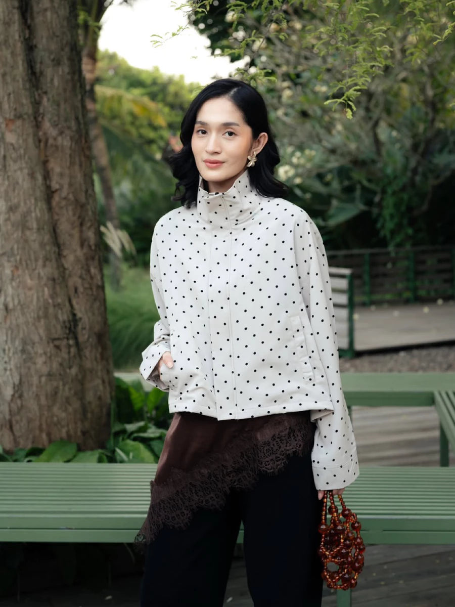 A woman standing outdoors in a park-like setting wearing a white long-sleeve mock-neck shirt by Haidee Orlin with black polka dots, a dark brown lace-trimmed wrap or skirt layer, black pants, and holding a red beaded handbag.