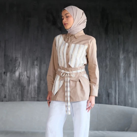 Woman wearing a beige blouse by Tunique with a striped tie and white pants against a dark textured wall.