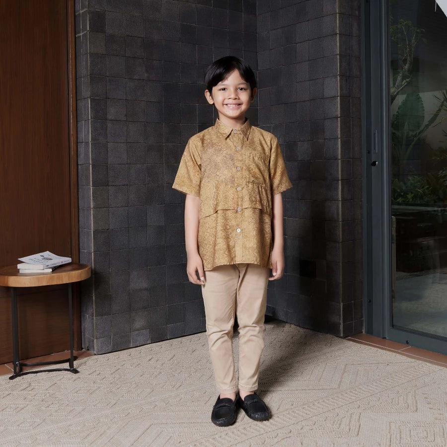 A boy wearing a patterned gold shirt by Jenna & Kaia and beige pants standing indoors against a dark wall.