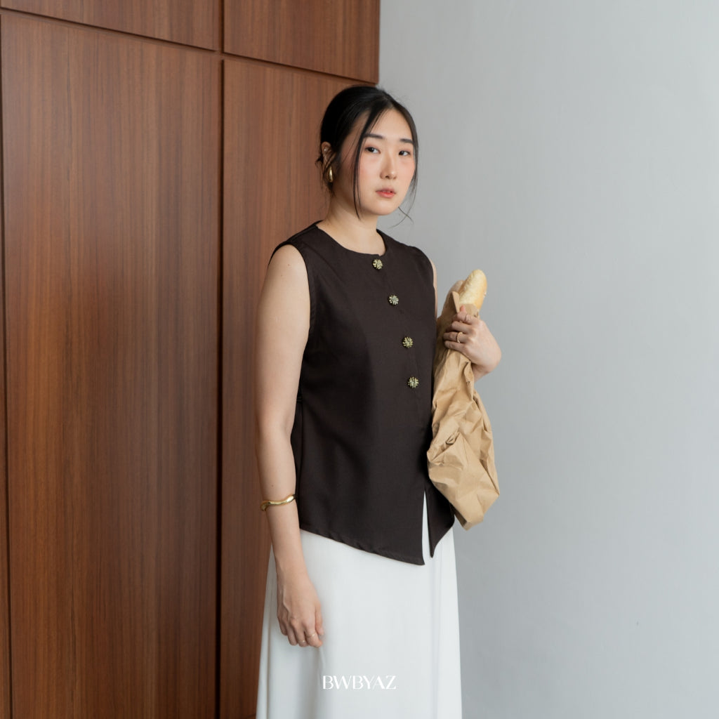 A person wearing a sleeveless deep bronze vest from BWBYAZ over a white skirt, standing against a wooden background.