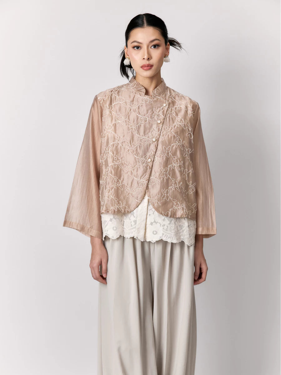 Woman wearing a beige lace outer by Studio Tui with a white lace overlay and beige pants on a light gray background