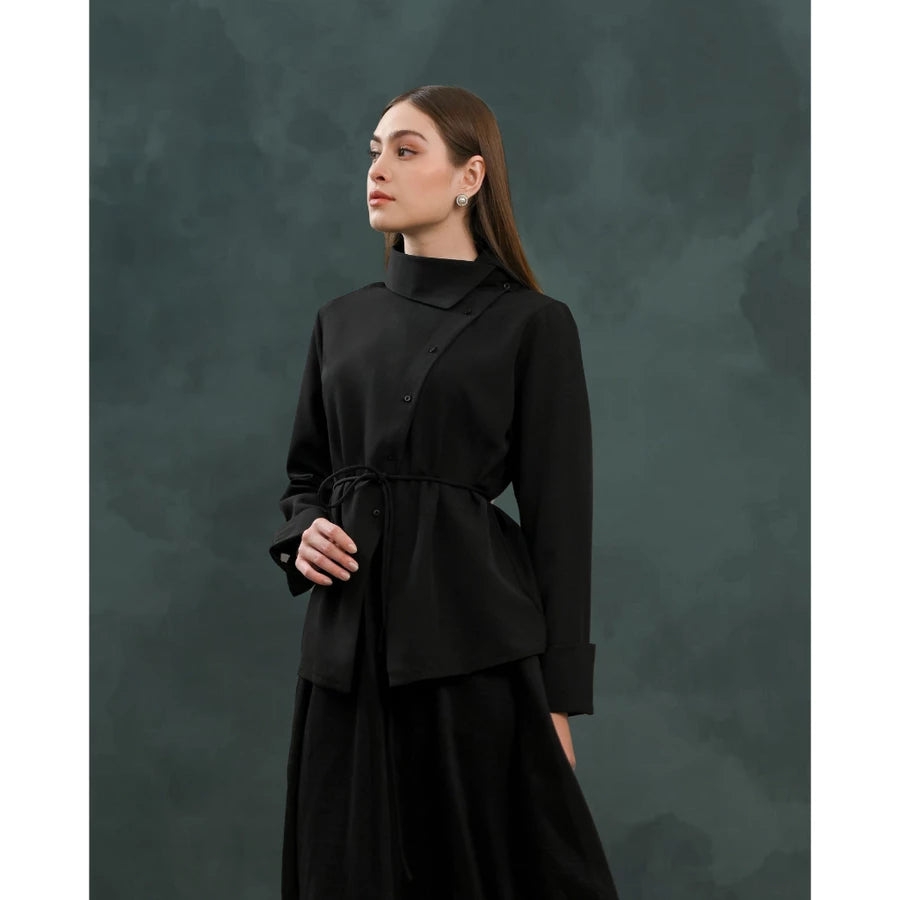 A woman modeling a black long-sleeve wrap-style blouse by Kasa Heritage with a drawstring waist, paired with a black skirt, set against a dark textured studio backdrop.