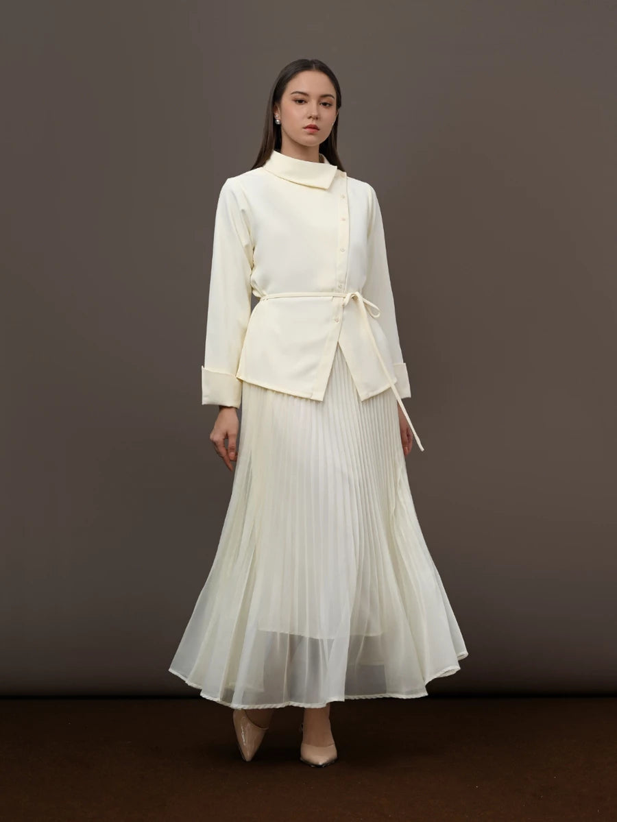 A model stands against a dark brown studio backdrop wearing a cream-colored two-piece outfit consisting of a long-sleeve wrap-style blouse by Kasa Heritage with a tie belt and a matching pleated maxi skirt.