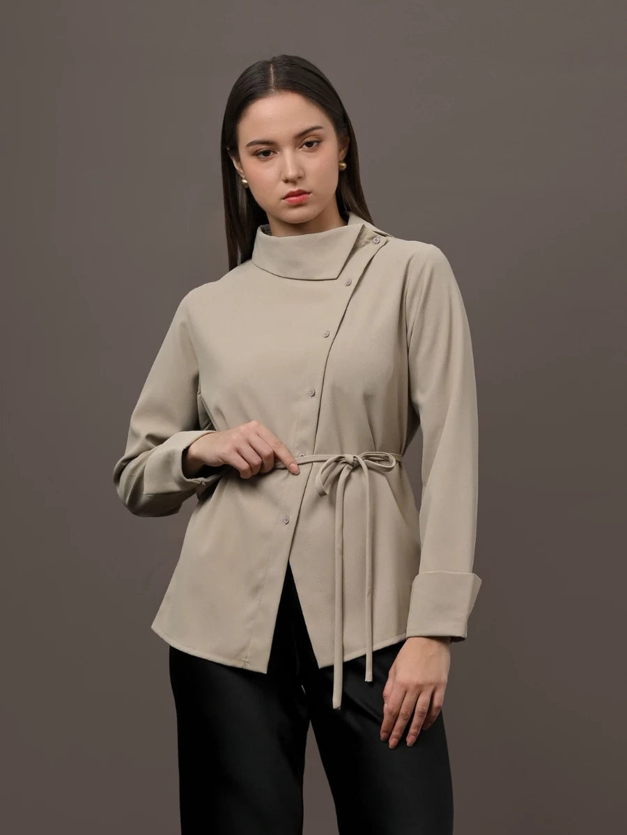 A woman modeling a beige long-sleeve wrap-style blouse by Kasa Heritage with a high mock neck and a drawstring tie at the waist, paired with black pants, against a solid dark gray studio background.