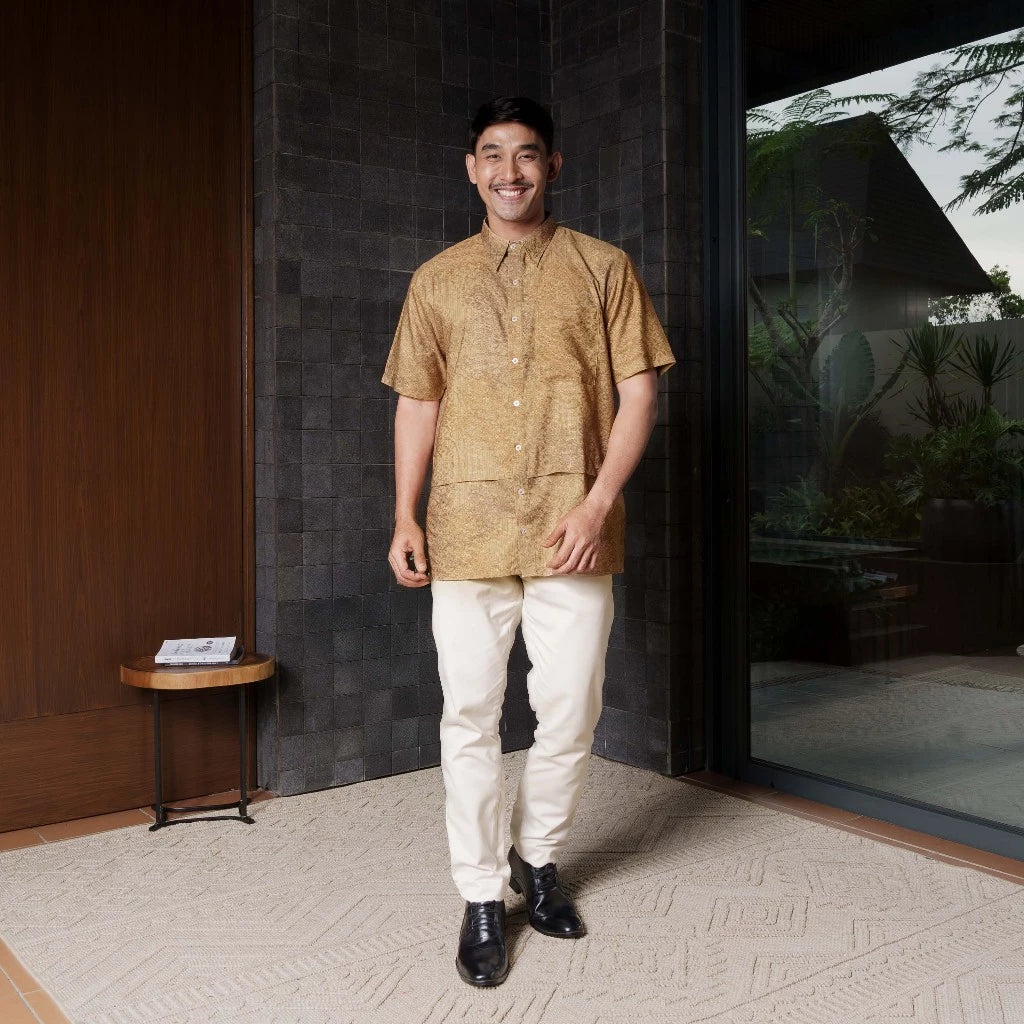 Man in a gold shirt by Jenna & Kaia and white pants standing in a modern indoor setting.