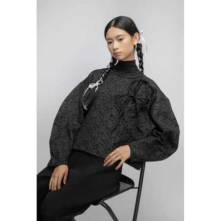 Woman wearing a black textured blouse by Yestoday with puffed sleeves and black pants, sitting on a chair against a light gray background.