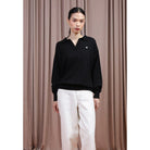 A model stands against a brown curtain backdrop wearing a black long-sleeve polo-style knit top by Hijabchic with a small white embroidered logo on the chest, paired with white pants.