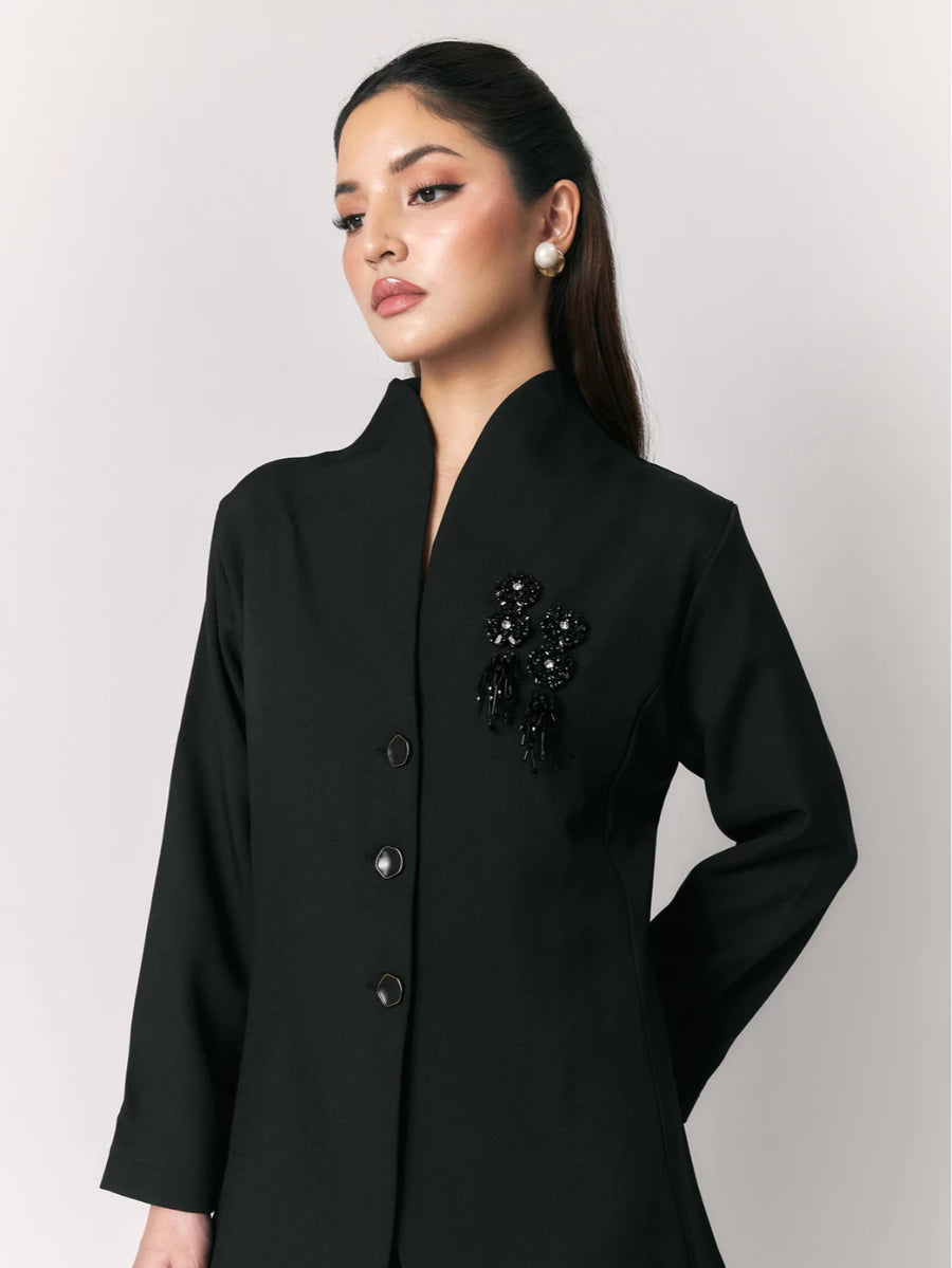 A woman wearing a black blazer by Studio Tui with a stand collar, three front buttons, and a black beaded embellishment on the left chest, photographed against a light gray studio background.
