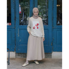 A woman wearing a beige top by Maima with a floral design on the front, paired with a long skirt and a headscarf, standing in front of a blue door.