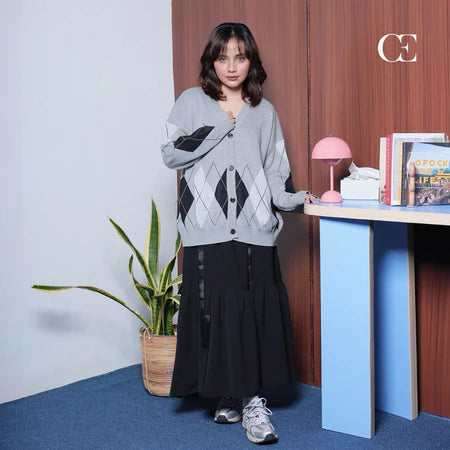 A person standing in a room wearing a grey fir cardigan by Casa Elana over a black top, paired with a long black skirt and white sneakers.