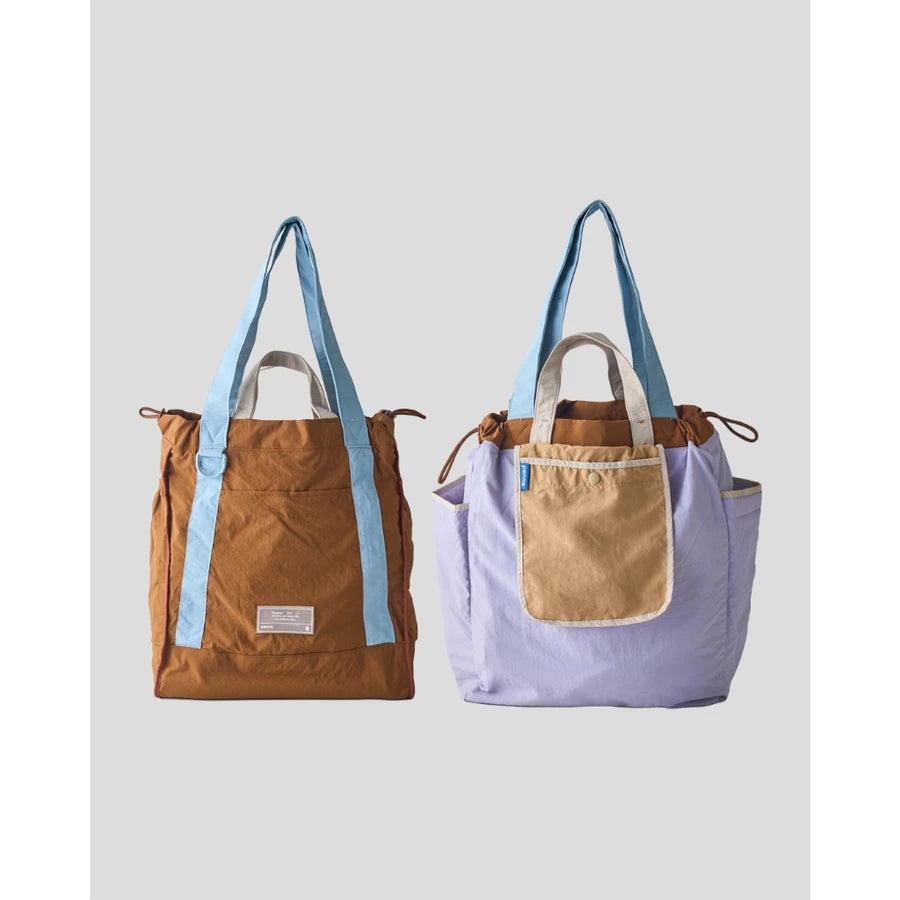 Reversible tote bag by Muhajir Project displayed in two sides, showing dual-color design with light blue and brown combination on one side and lilac with light brown pocket on the other. This versatile women’s bag features long shoulder straps, side drawstring details, and a functional front pocket, highlighting its practical and stylish two-in-one design.