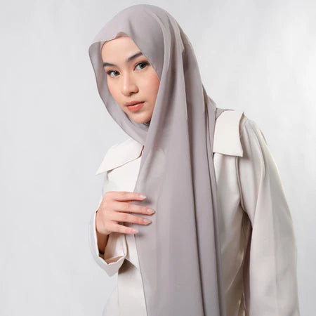 Woman wearing a light gray pashmina hijab by Tantri Namirah Official and white outfit on a plain background