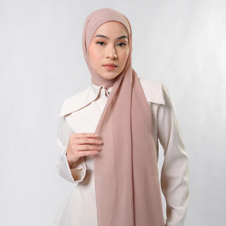 Woman wearing a light pink pashmina hijab by Tantri Namirah Official and white shirt against a plain background