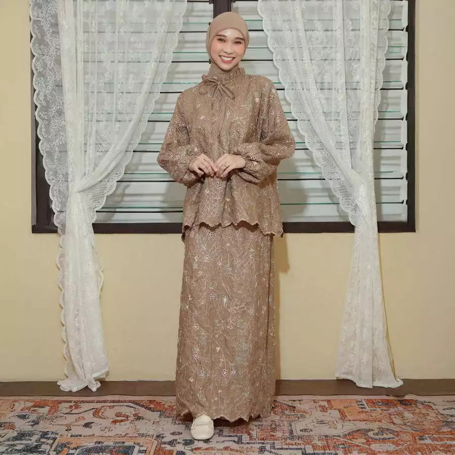 Woman in a beige traditional outfit by Lozy Hijab standing in front of a window with white curtains.