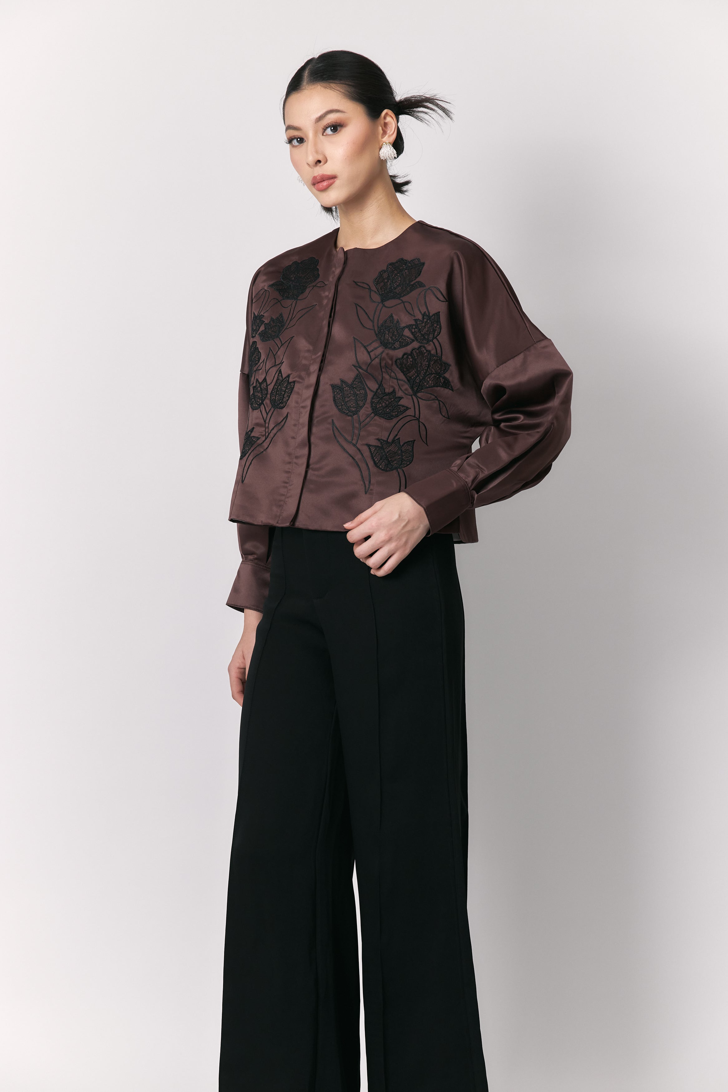 Person wearing a brown outer with floral embroidery by Studio Tui and black pants on a white background