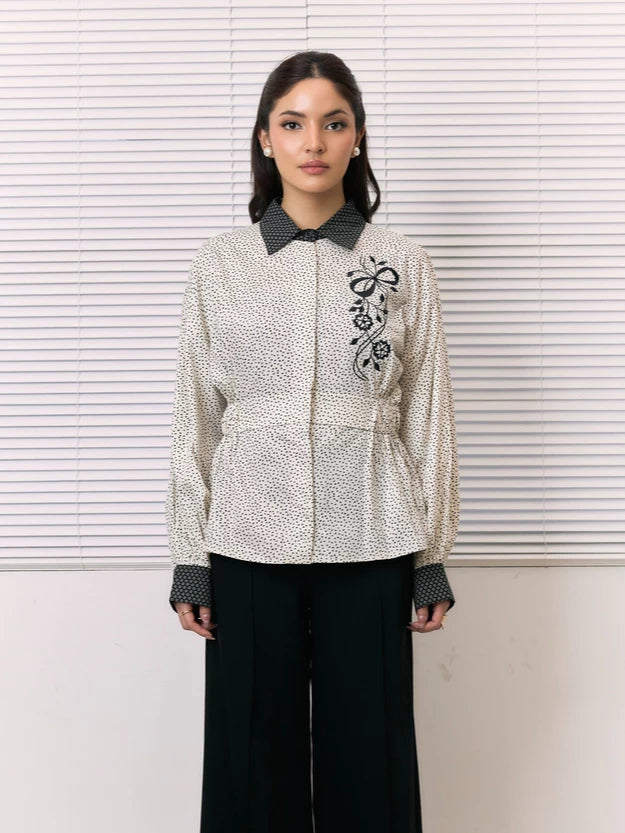 A woman stands in front of white window blinds wearing a cream-colored textured shirt by Studio Tui with black floral embroidery on the chest, a black patterned collar and cuffs, and black wide-leg pants.