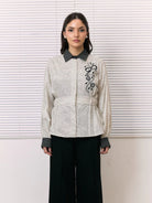 A woman stands in front of white window blinds wearing a cream-colored textured shirt by Studio Tui with black floral embroidery on the chest, a black patterned collar and cuffs, and black wide-leg pants.