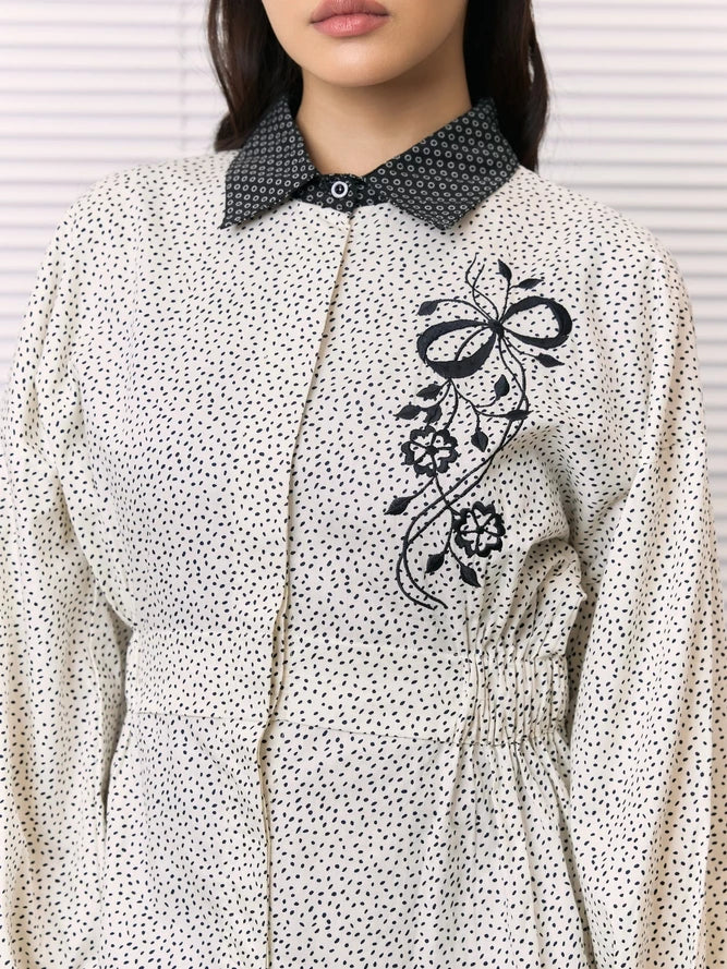 A close-up of a woman wearing a cream-colored long-sleeve shirt by Studio Tui with a black micro-dot pattern, a contrasting black patterned collar, and a black floral embroidery on the chest.