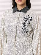 A close-up of a woman wearing a cream-colored long-sleeve shirt by Studio Tui with a black micro-dot pattern, a contrasting black patterned collar, and a black floral embroidery on the chest.