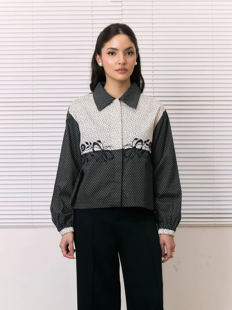 A woman stands in front of white window blinds wearing a cropped long-sleeve shirt by Studio Tui with a black-and-white patterned body, contrasting solid black collar and sleeves, and black pants.