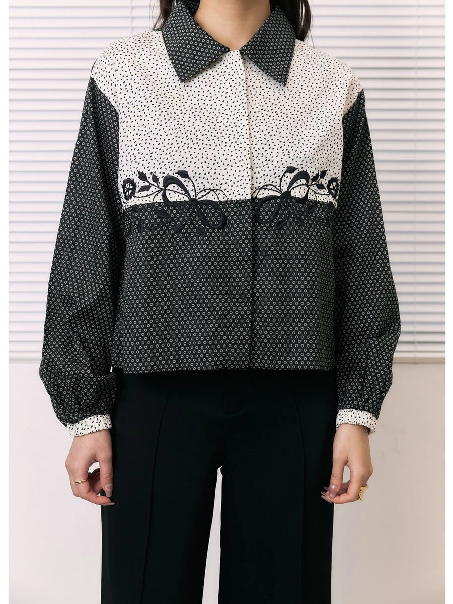 A woman wearing a long-sleeve shirt by Studio Tui with a color-blocked design: a cream and black micro-dot pattern on the upper chest and shoulders, solid black sleeves, and a black-and-white geometric pattern across the lower half. She is also wearing black pants.