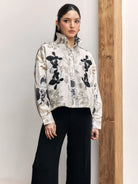 A woman modeling a cropped, long-sleeve high-neck shirt by Studio Tui with a black, gray, and white floral pattern on a cream background, paired with black wide-leg pants, standing next to a wooden pillar.