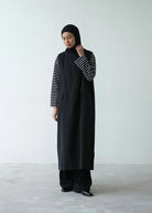 A person wearing a sleeveless dress by Rashawl paired with long sleeves shirt standing against a plain background.