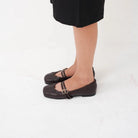A person wearing dark brown leather Mary Jane flatshoes by Cekhas with double straps and small gold buckle details, paired with a black skirt, shown from the knees down on a white studio background.