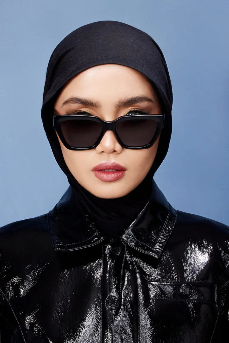 A person wearing a black headscarf and black jacket, with the sunglasses from HSF Eyewear covering the eyes. The sunglasses have a black frame and black lenses.