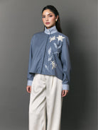 A model wearing a dusty blue cropped shirt by Studio Tui with white floral embroidery on the chest, paired with cream wide-leg trousers, against a solid gray studio background.